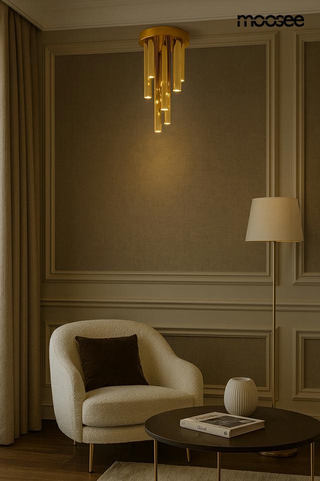 Lampa sufitowa Organo z dziewięcioma diodami LED, moc 10W, barwa światła 3000K. Złota, wisząca konstrukcja z wydłużonymi elementami, umieszczona w eleganckim wnętrzu z jasnymi zasłonami i stylowym fotelem.