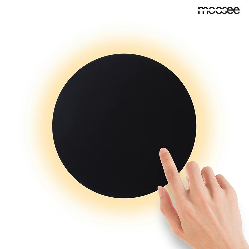 Czarny kinkiet LED Shadow Sun 25 z jednolitą, okrągłą formą, emitujący ciepłe światło wokół krawędzi. Produkt wisi na białym tle, z ręką dotykającą powierzchni lampy, co sugeruje interaktywność.