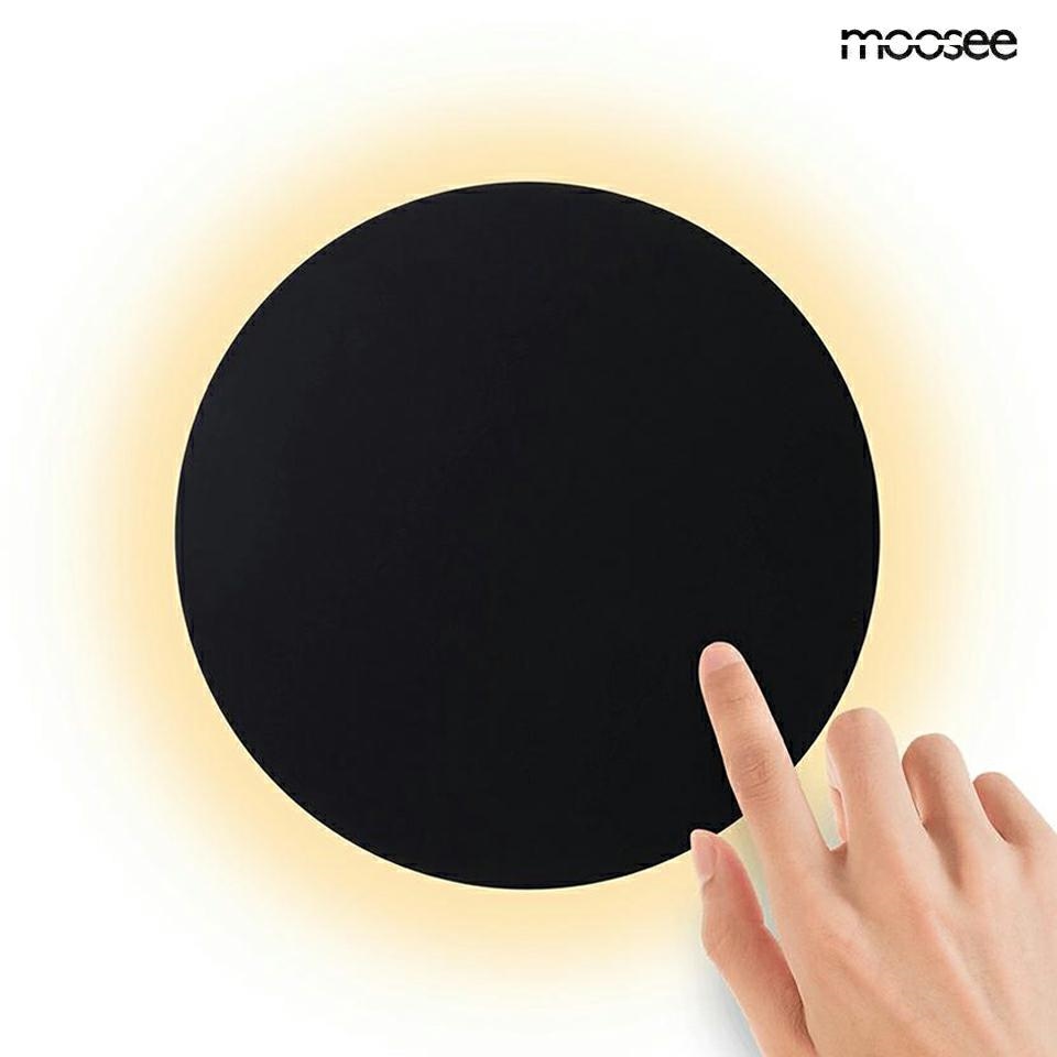 Kinkiet LED Shadow Sun 30 w kolorze czarnym, o okrągłym kształcie. Widoczna ręka dotykająca powierzchni lampy, która emituje ciepłe światło (3000K). Proste tło, podkreślające minimalistyczny design.