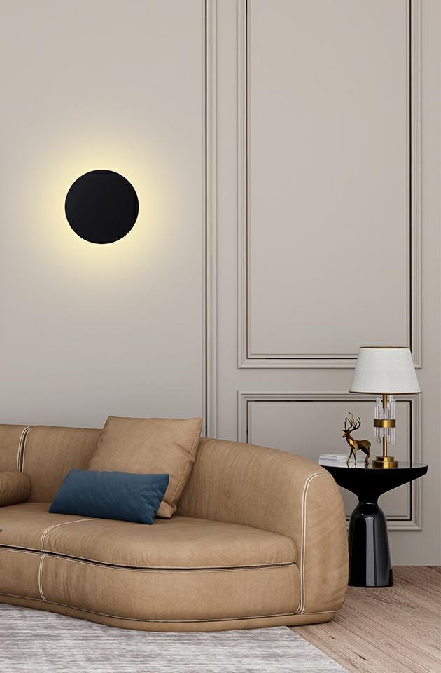 Kinkiet LED Shadow Sun 30, 8W, 3000K w kolorze czarnym. Okrągły kształt z subtelnym podświetleniem, zamontowany na ścianie nad sofą w nowoczesnym wnętrzu. Tło w jasnym kolorze z eleganckim wykończeniem.