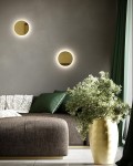 Kinkiet LED Shadow Sun 25 w kolorze złotym, emitujący ciepłe światło 3000K. Zamontowany na ścianie, w kształcie okręgu. Tło to szara ściana z elementami dekoracyjnymi, takimi jak ciemna sofa i roślina w złotym wazonie.