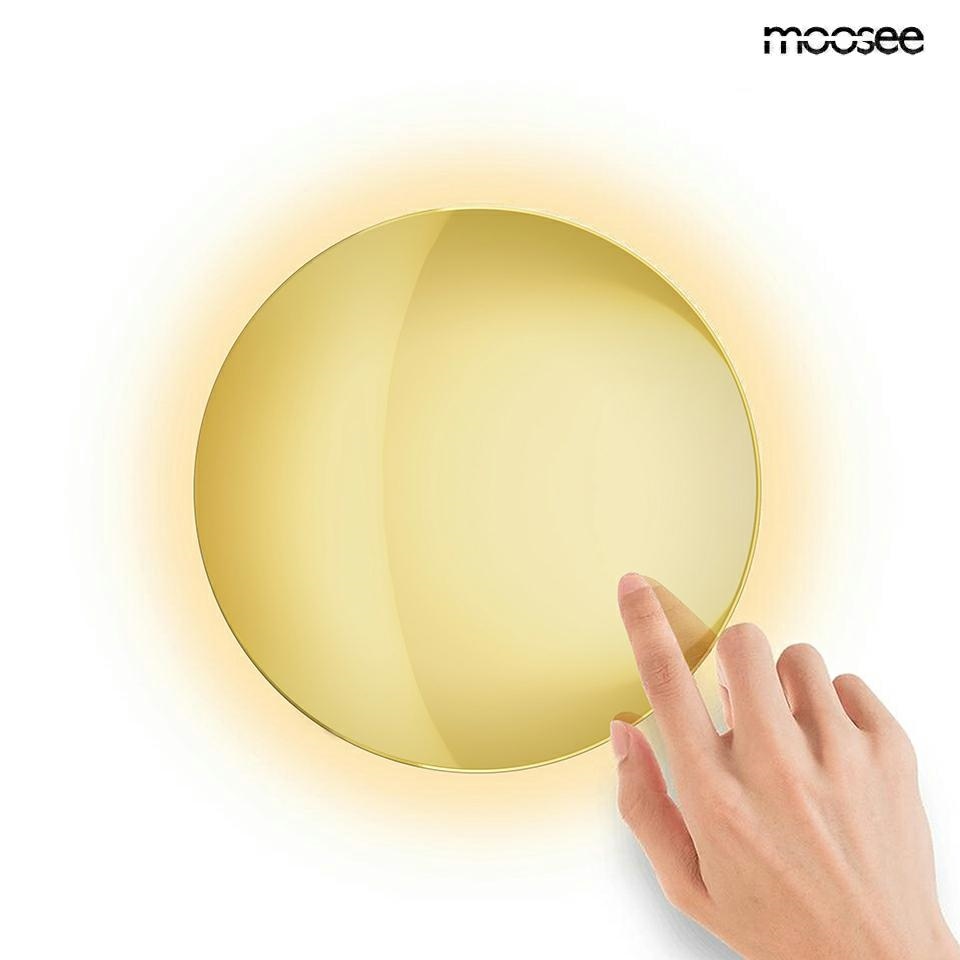 Kinkiet LED Shadow Sun 25 o okrągłym, złotym kloszu, emitującym ciepłe światło 3000K. Produkt dotykowy, widać rękę dotykającą powierzchni. Tło białe, podkreślające elegancję lampy. Wymiary i szczegóły konstrukcyjne nie są widoczne.