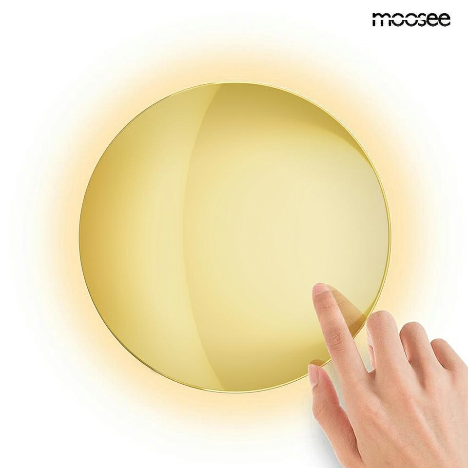 Kinkiet LED Shadow Sun 30 w kolorze złotym o mocy 8W i temperaturze barwowej 3000K. Okrągły kształt, gładka, błyszcząca powierzchnia. W ręce osoba dotyka lampy, co sugeruje jej interaktywną funkcję. Tło jasne, podkreślające elegancję produktu.