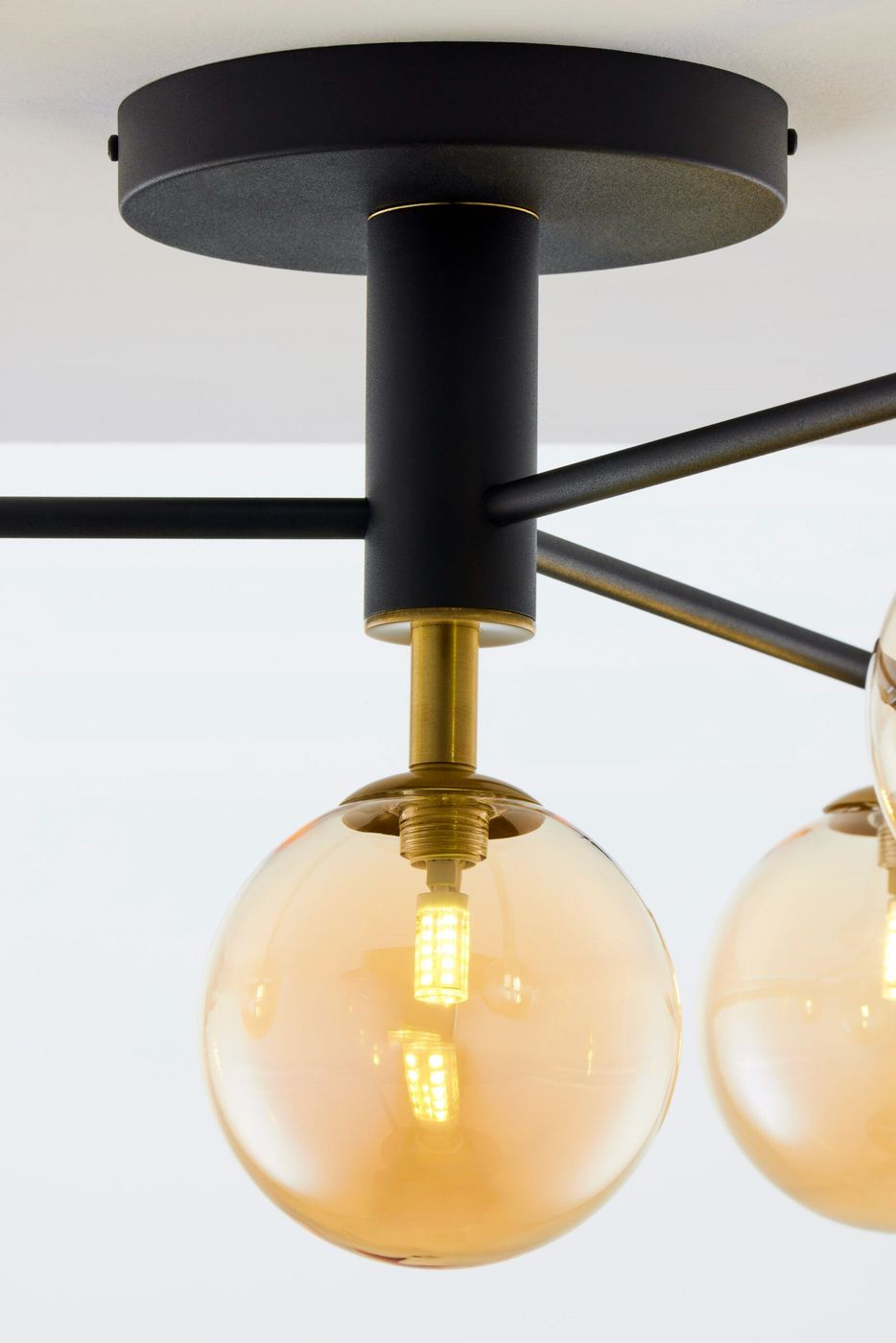 Lampa sufitowa Figaro w kolorze czarnym z bursztynowymi kloszami. Posiada cztery źródła światła G9. Ujęcie z boku, ukazujące detal mocowania i połyskujący klosz. Metalowa konstrukcja z elementami w kolorze złotym, na białym tle.