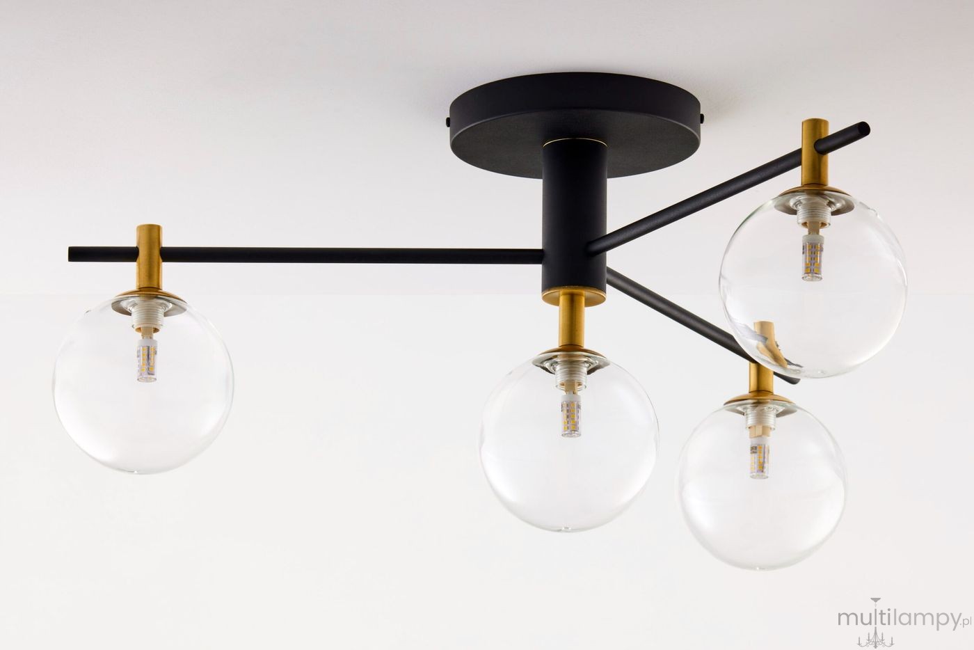 Lampa sufitowa Figaro z czarnym metalowym stelażem i transparentnymi szklanymi kloszami. Posiada cztery źródła światła G9, rozmieszczone na poziomej metalowej rurze, z elementami w kolorze złotym. Ujęcie z dołu na zamontowaną lampę.