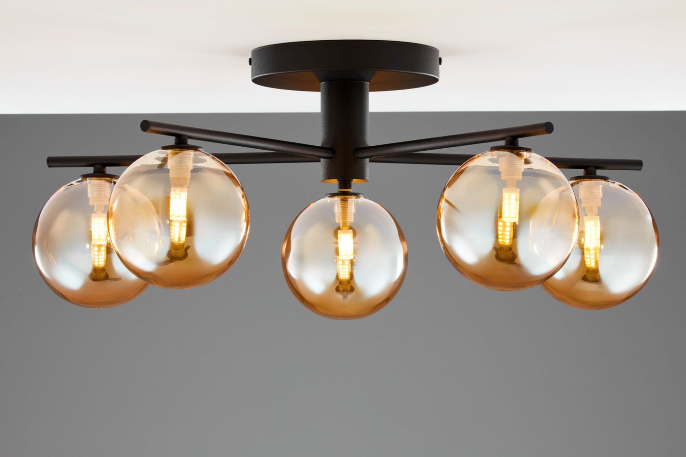 Lampa sufitowa Prato w kolorze czarnym z pięcioma bursztynowymi kloszami G9. Klosze w kształcie kul, zamontowane na metalowej konstrukcji, z widocznymi żarówkami. Ujęcie z dołu, ukazujące detal i kształt lampy. Tło w odcieniach szarości.