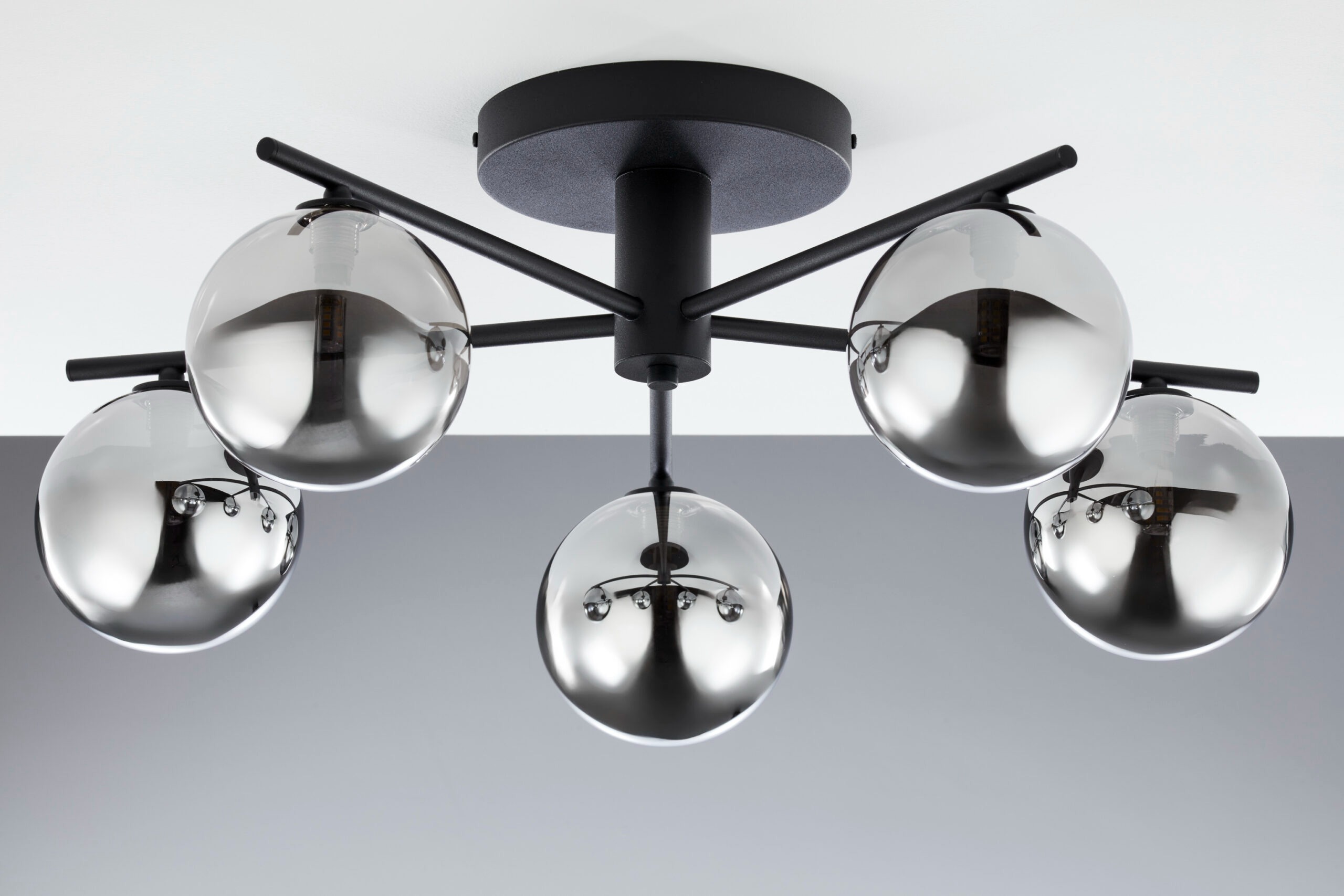 Lampa sufitowa Prato w kolorze czarnym, z pięcioma dymnymi kulami G9, zamontowana na okrągłej podstawie. Kule mają metaliczne wykończenie, a konstrukcja jest minimalistyczna, z poziomymi ramionami. Ujęcie z dołu, pokazujące detale.