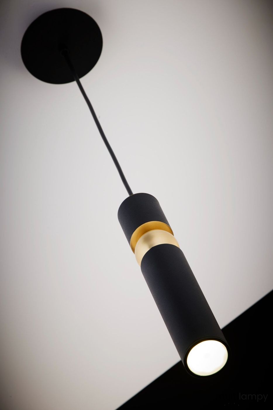 Czarna lampa wisząca Alas z jedną oprawą GU10, o cylindrycznym kształcie z matowym wykończeniem. Posiada złotą dekorację w środkowej części. Ujęcie z dołu, zawieszona na cienkim przewodzie. Tło białe, podkreślające elegancki design.