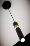 Czarna lampa wisząca Alas z jedną oprawą GU10, o cylindrycznym kształcie z matowym wykończeniem. Posiada złotą dekorację w środkowej części. Ujęcie z dołu, zawieszona na cienkim przewodzie. Tło białe, podkreślające elegancki design.
