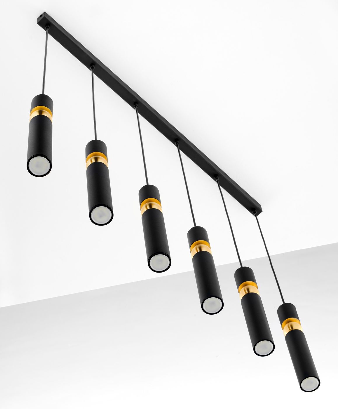 Lampa wisząca Alas z sześcioma źródłami światła GU10. Czarna obudowa z metalowym wykończeniem w kolorze złotym. Ułożona poziomo, zawieszona na cienkich przewodach, z cylindrycznymi kloszami w dół. Prosty, nowoczesny design.