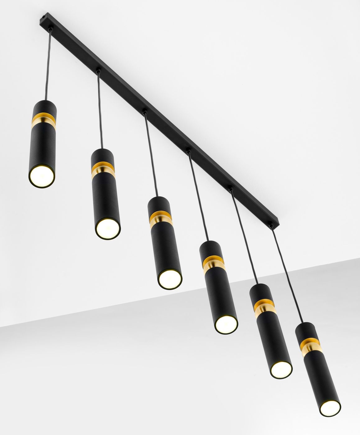 Lampa wisząca Alas z sześcioma źródłami światła GU10, czarna z metalowymi złotymi akcentami. Wisi na prostokątnej podstawie, z lampami w formie cylindrów, zawieszonymi na cienkich linkach. Tło białe, minimalistyczne.