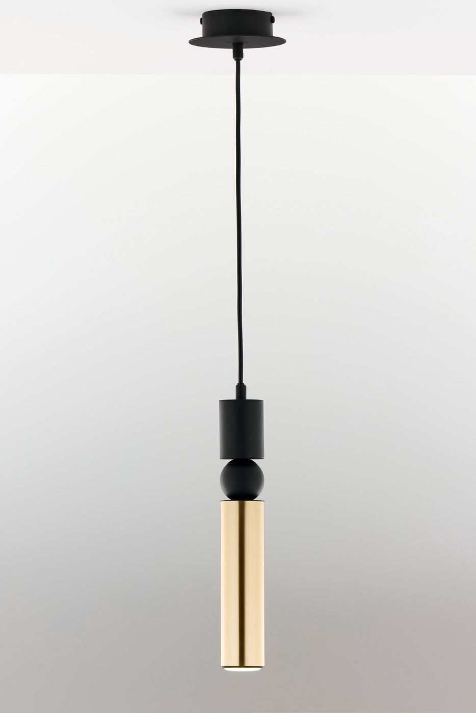 Lampa wisząca Alas z jednym miejscem na żarówkę GU10, w kolorze czarnym i złotym. Posiada cylindryczny, złoty klosz oraz czarną podstawkę i kabel. Ujęcie frontalne, zawieszona z sufitu na prostym tle.