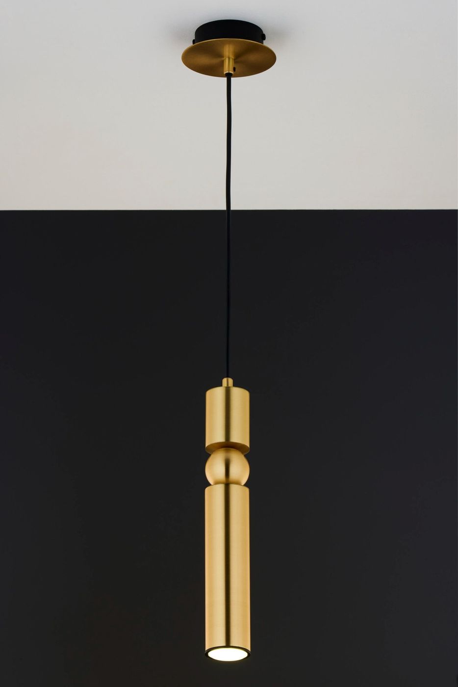 Lampa wisząca Alas w kolorze złotym, z jednym źródłem światła GU10. Posiada cylindryczny kształt z dekoracyjnymi elementami, zawieszona na czarnym przewodzie. Ujęcie frontalne, na jednolitym ciemnym tle.