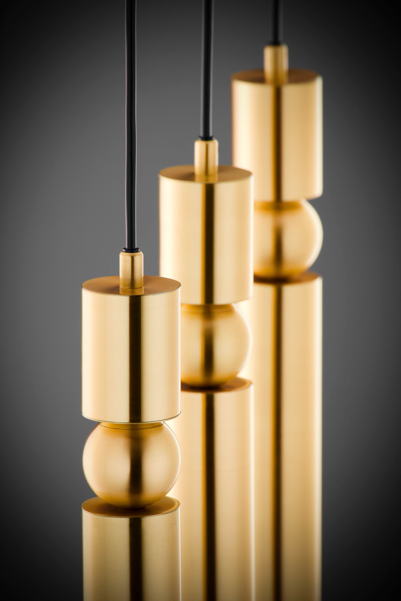 Złota lampa wisząca Alas z trzema źródłami światła GU10. Kształt cylindryczny z zaokrąglonymi detalami, zawieszona na cienkich czarnych przewodach. Powierzchnia gładka, błyszcząca, na ciemnym tle, podkreślająca elegancki design.