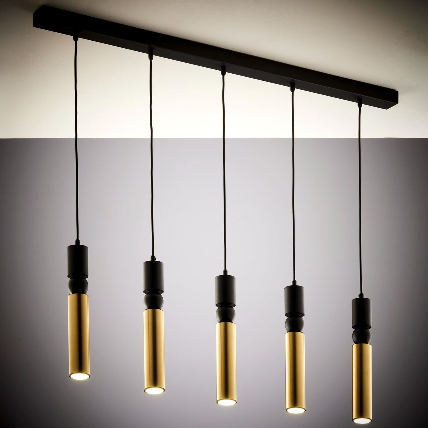 Lampa wisząca Alas z pięcioma źródłami światła GU10. Czarna konstrukcja z wiszącymi cylindrycznymi kloszami w kolorze złotym, zawieszona na czarnym przewodzie. Ujęcie z góry, na jednolitym tle.