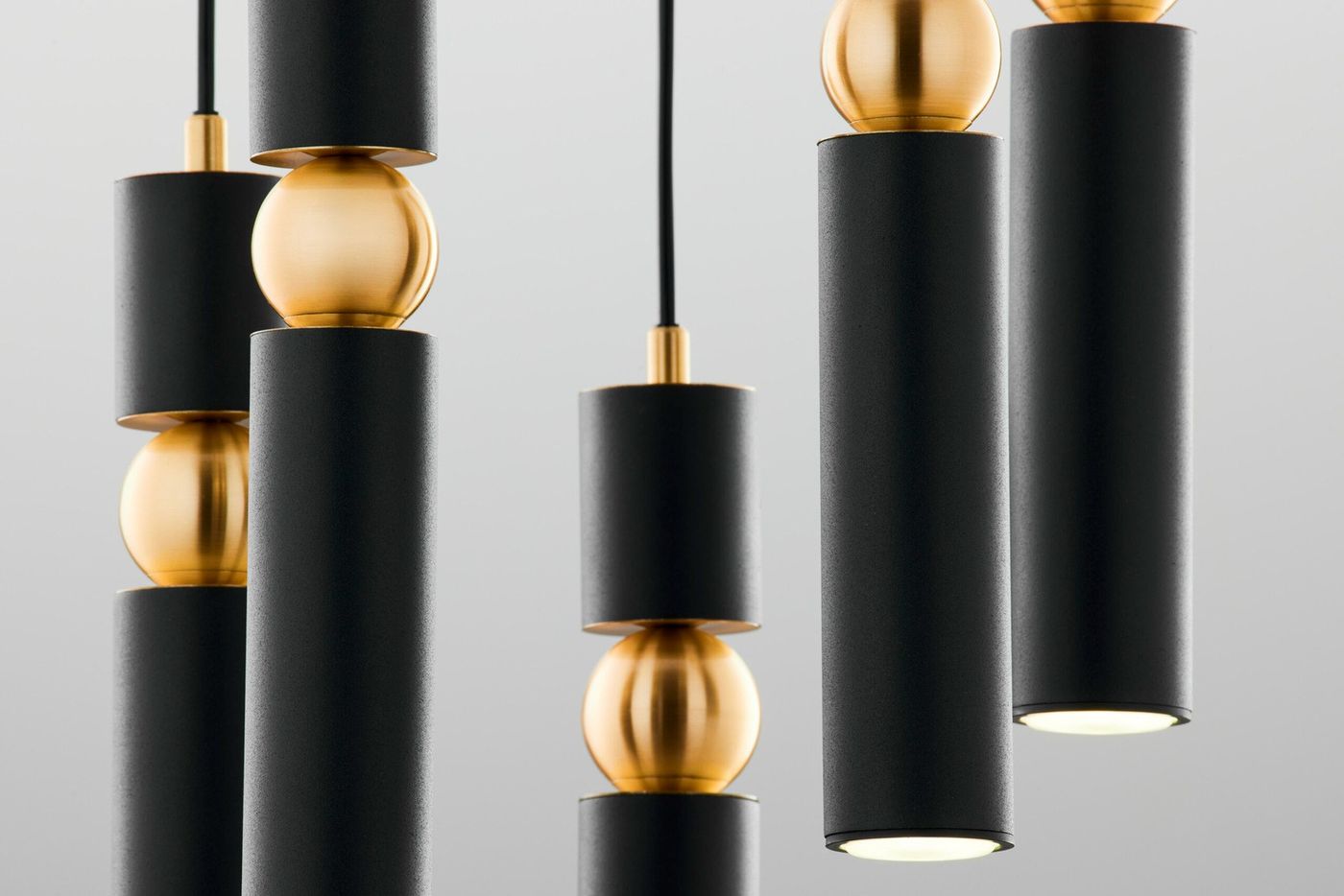 Lampa wisząca Alas w kolorze czarnym z pięcioma oprawami GU10. Składa się z cylindrycznych elementów w matowym wykończeniu oraz metalowych kul w kolorze złotym. Produkt zawieszony na cienkim kablu, ukazujący nowoczesny design.