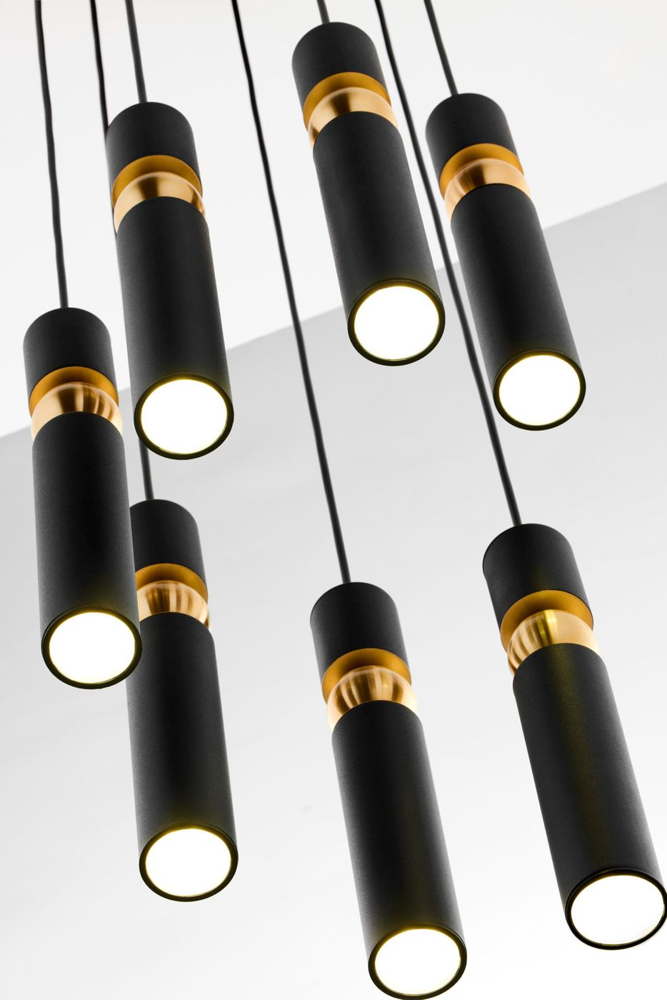 Lampa wisząca Alas z 7 oprawami GU10, w kolorze czarnym z elementami złotymi. Proste, cylindryczne kształty zwisają na czarnych przewodach. Ujęcie z bliska, pod kątem, ukazujące detale wykończenia oraz źródła światła.