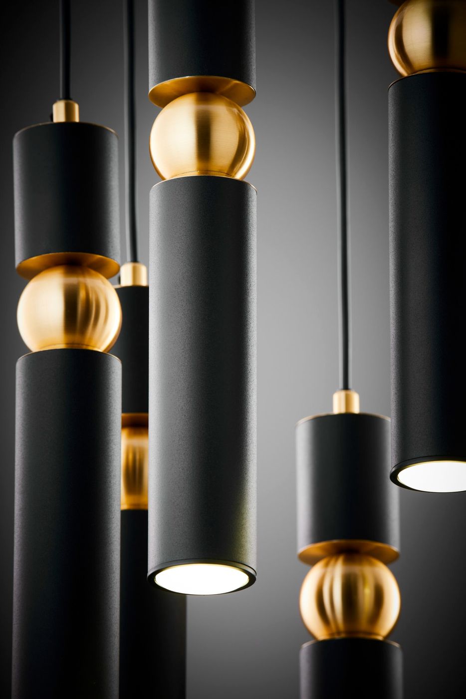 Lampa wisząca Alas, czarna, z siedmioma cylindrycznymi kloszami, ozdobiona złotymi kulami. Ujęcie z bliska, ukazujące detale konstrukcji i wykończenia. Klosze emitują światło z dołu, zawieszone na cienkich linkach.