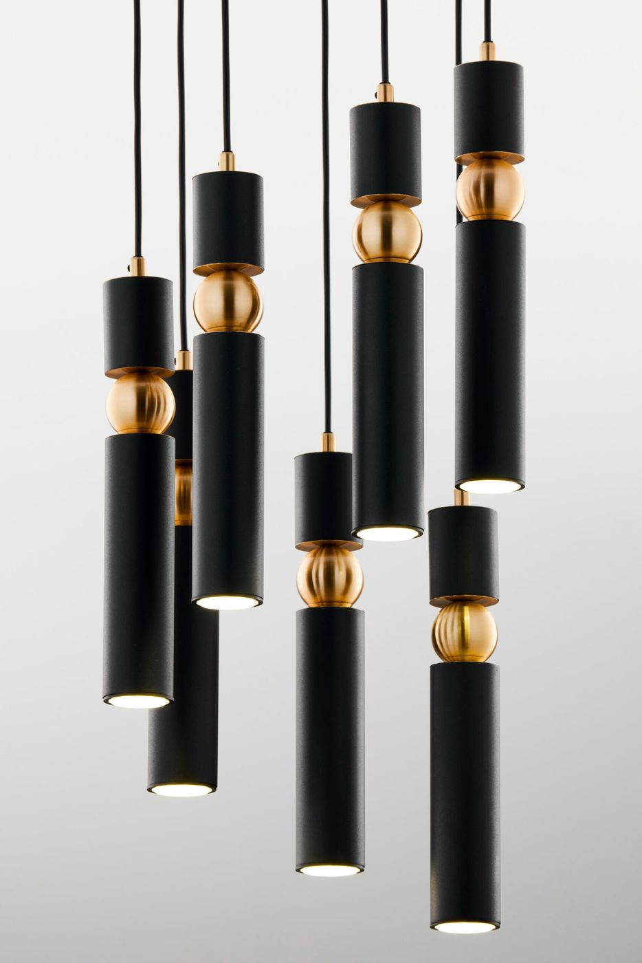 Czarna lampa wisząca Alas z siedmioma źródłami światła GU10. Elementy metalowe w kolorze złotym, cylindryczny kształt, różne długości zwisów. Lampa zawieszona na czarnych przewodach, minimalistyczny design. Tło jasne, jednolite.