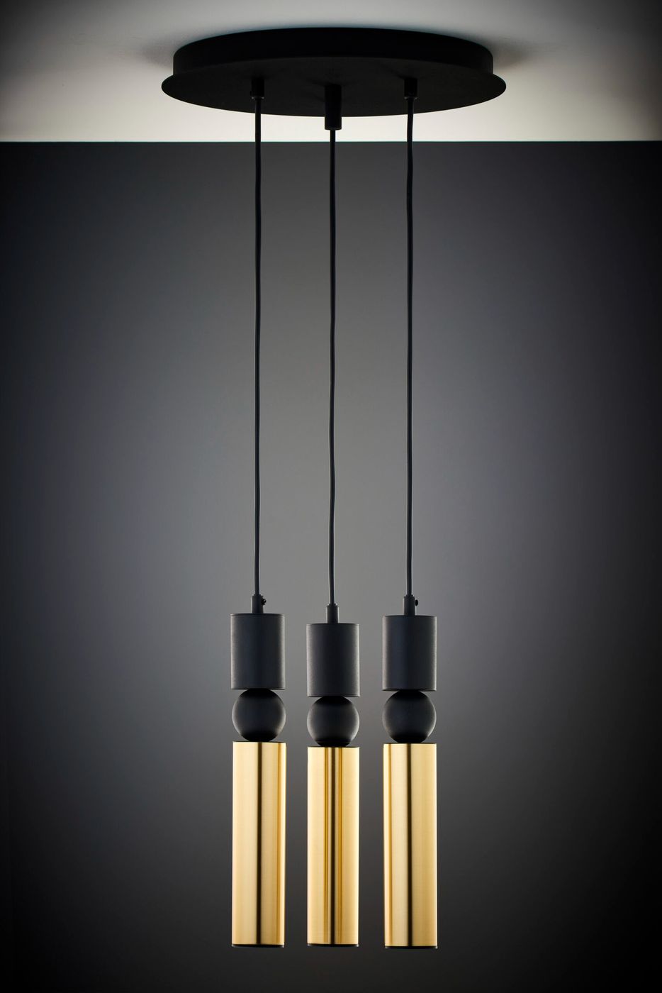 Lampa wisząca Alas z trzema źródłami światła GU10, w kolorze czarnym i złotym. Produkt z podwieszeniem na czarnych przewodach, składający się z trzech cylindrycznych elementów w kolorze złotym, zawieszonych na okrągłej podstawie. Tło w ciemnym odcieniu.