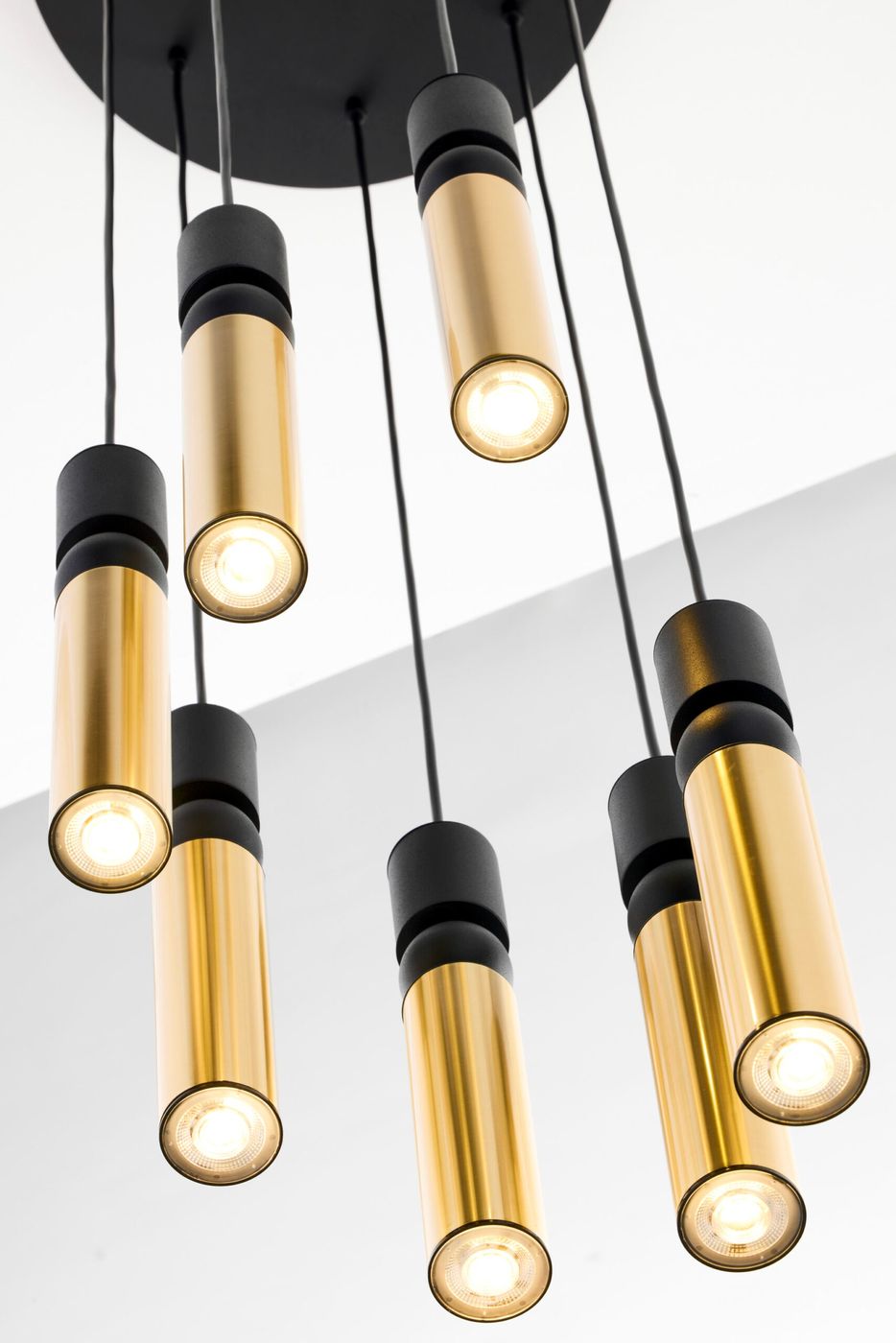 Lampa wisząca Alas z siedmioma źródłami światła GU10, złożona z cylindrycznych elementów w czarnym i złotym kolorze. Zawieszona na cienkich przewodach, montowana do sufitu, w nowoczesnym stylu. Idealna do aranżacji wnętrz.