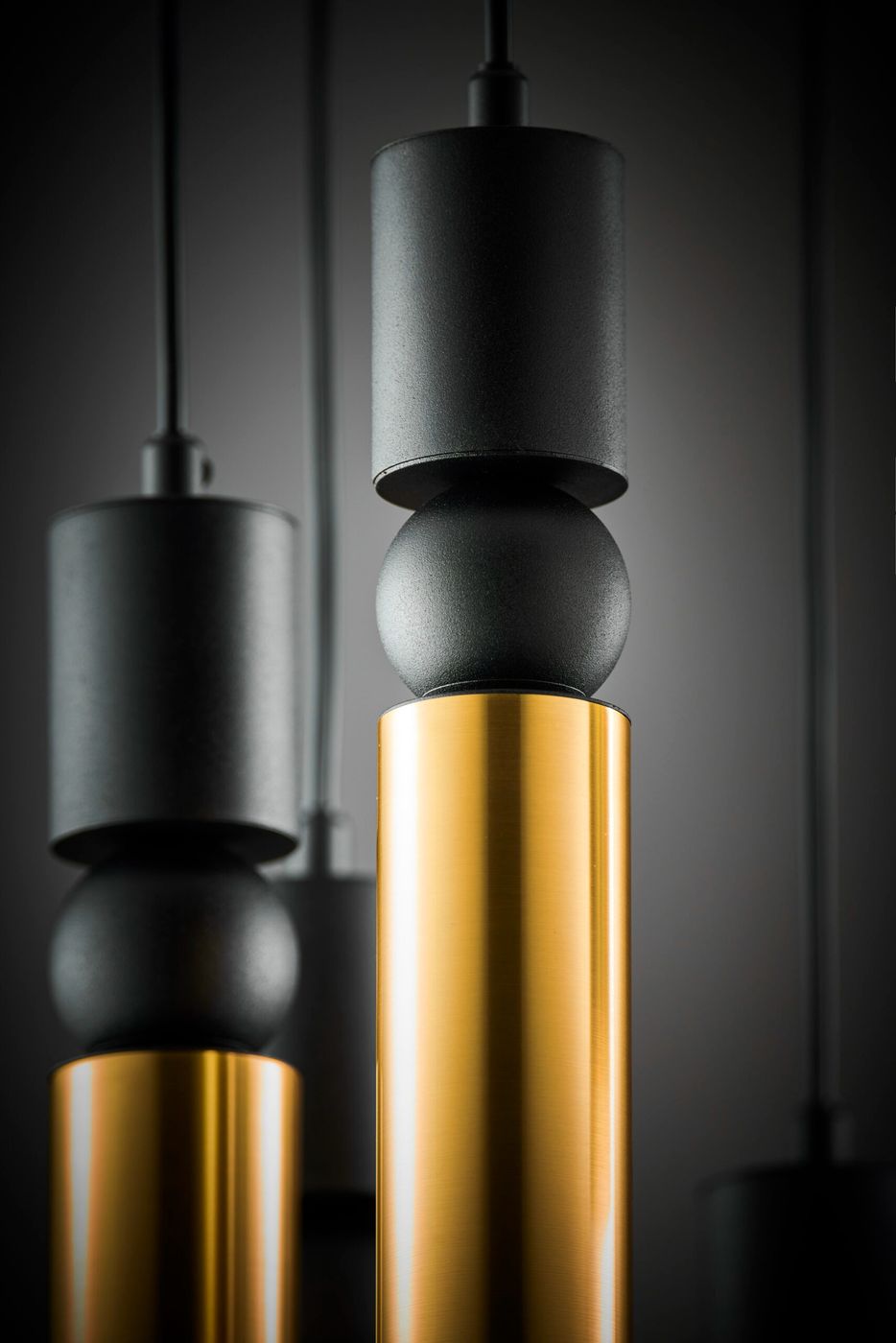 Lampa wisząca Alas w kolorze czarnym i złotym, z siedmioma miejscami na żarówki GU10. Widok z bliska ukazuje cylindryczne elementy: górny czarny klosz, kulistą część i złotą podstawę. Tło ciemne, co podkreśla elegancję lampy.