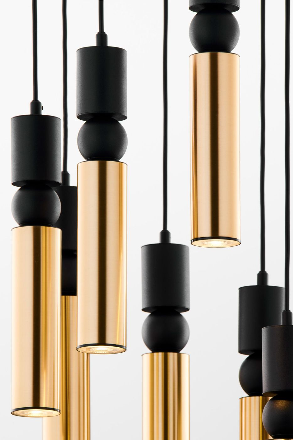 Lampa wisząca Alas z siedmioma źródłami światła GU10, w kolorze czarnym i złotym. Ujęcie z bliska, pokazujące cylindryczne, błyszczące elementy w złotym kolorze oraz matowe, czarne akcenty. Wisi na czarnych przewodach na jasnym tle.
