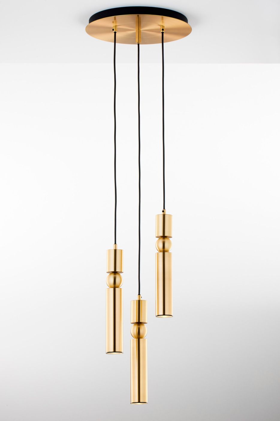 Lampa wisząca Alas z trzema źródłami światła GU10 w kolorze złotym. Produkt składa się z trzech cylindrycznych elementów w różnych wysokościach, zawieszonych na czarnych przewodach. Montaż na okrągłym, złotym stropie. Tło jasne, minimalistyczne.