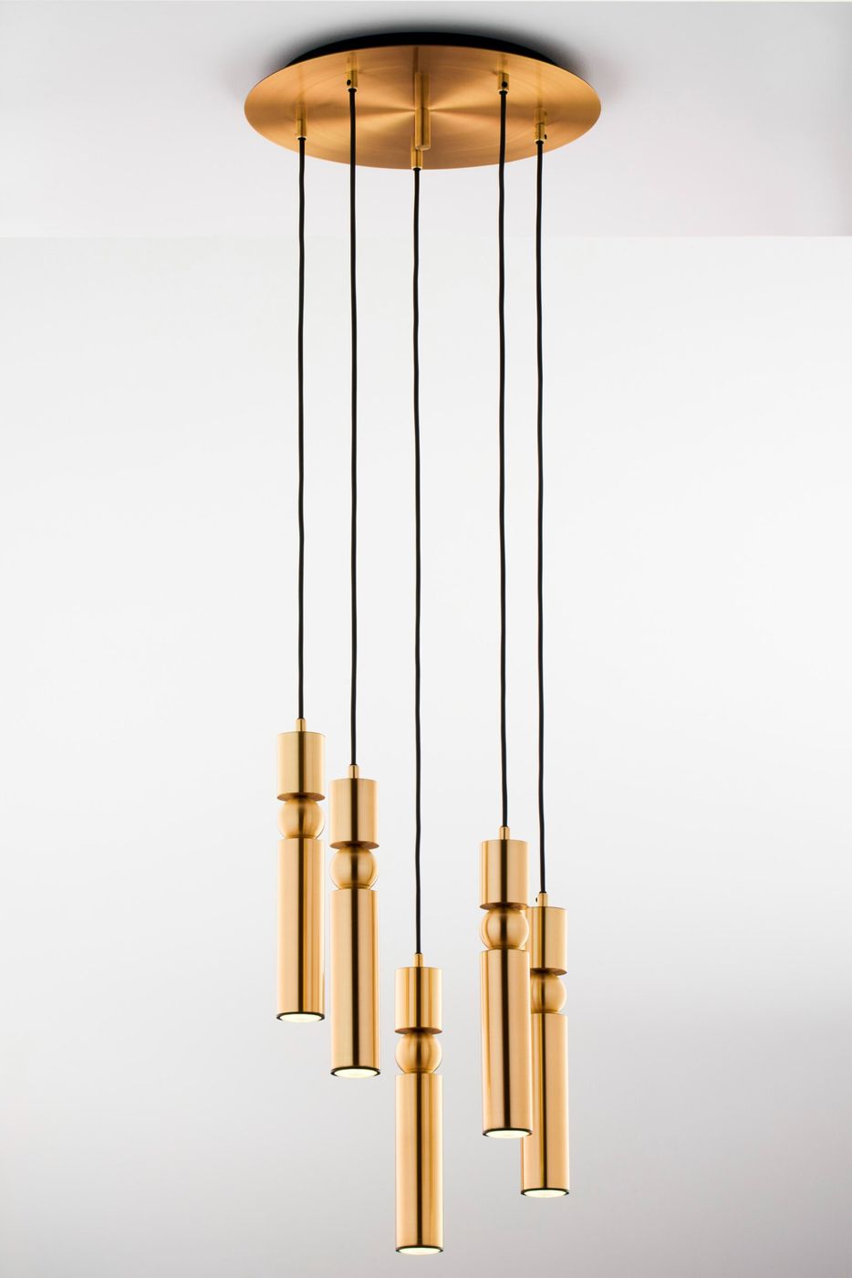 Lampa wisząca Alas z pięcioma oprawami GU10, w kolorze złotym. Posiada cylindryczne klosze o różnej wysokości zawieszone na czarnych kablach. Montaż sufitowy na okrągłej, złotej podstawie. Tło białe, minimalistyczne.