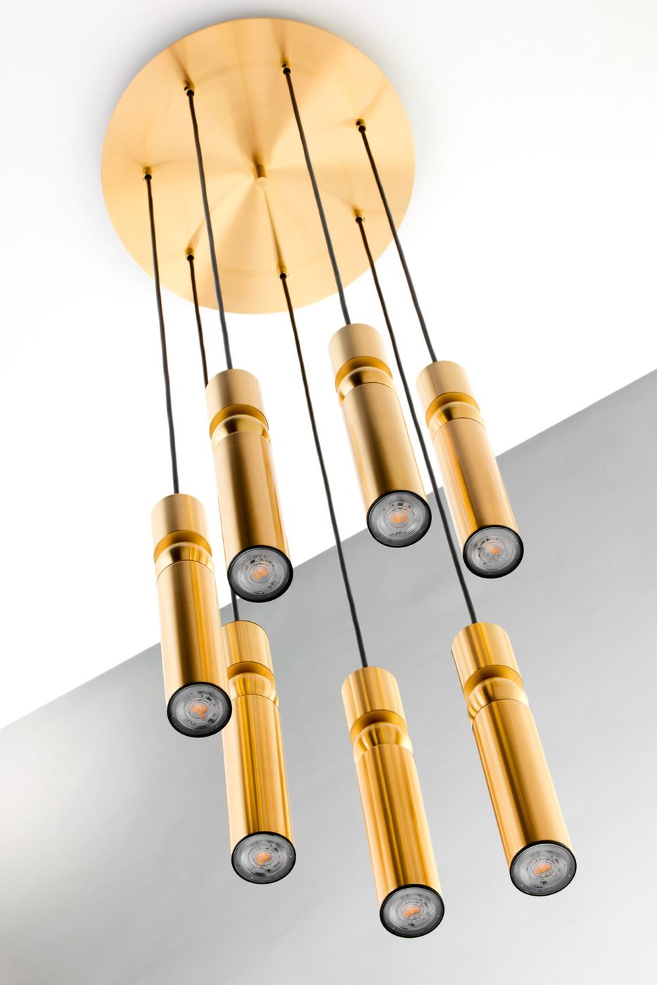 Lampa wisząca Alas 7xGU10 w kolorze złotym. Sześć cylindrycznych kloszy zawieszonych na czarnych linkach z centralnym złotym mocowaniem. Klosze mają czarne końcówki i przezroczyste szkło, emitują światło. Ujęcie z dołu, na jasnym tle.