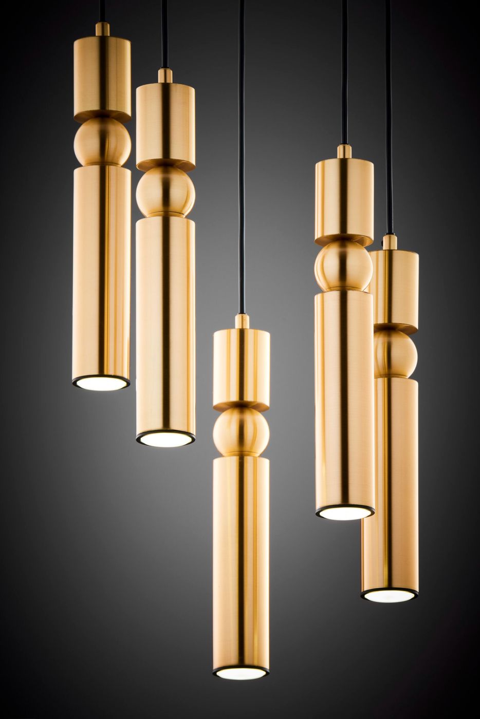 Lampa wisząca Alas z pięcioma źródłami światła GU10, w kolorze złotym. Składa się z cylindrycznych elementów o różnych wysokościach, zawieszonych na czarnych kablach. Ujęcie z przodu, na ciemnym tle.