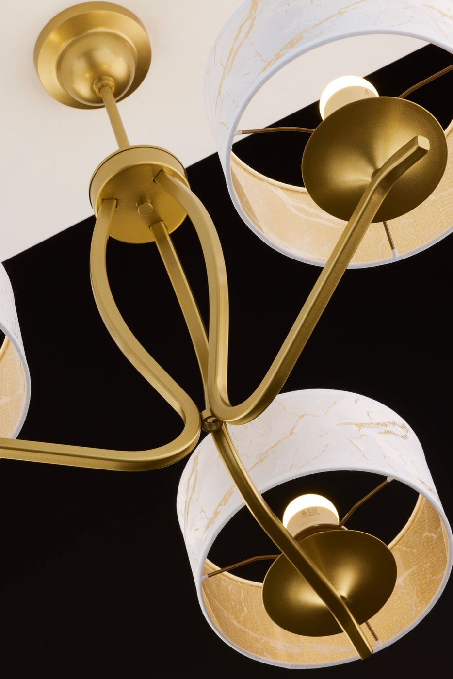 Kaszmir żyrandol w kolorze złotym z białymi kloszami, posiada trzy źródła światła E27. Wzory w złotym odcieniu na kloszach, metalowa konstrukcja, widok z dołu, z widocznymi detalami mocowania i żarówek.