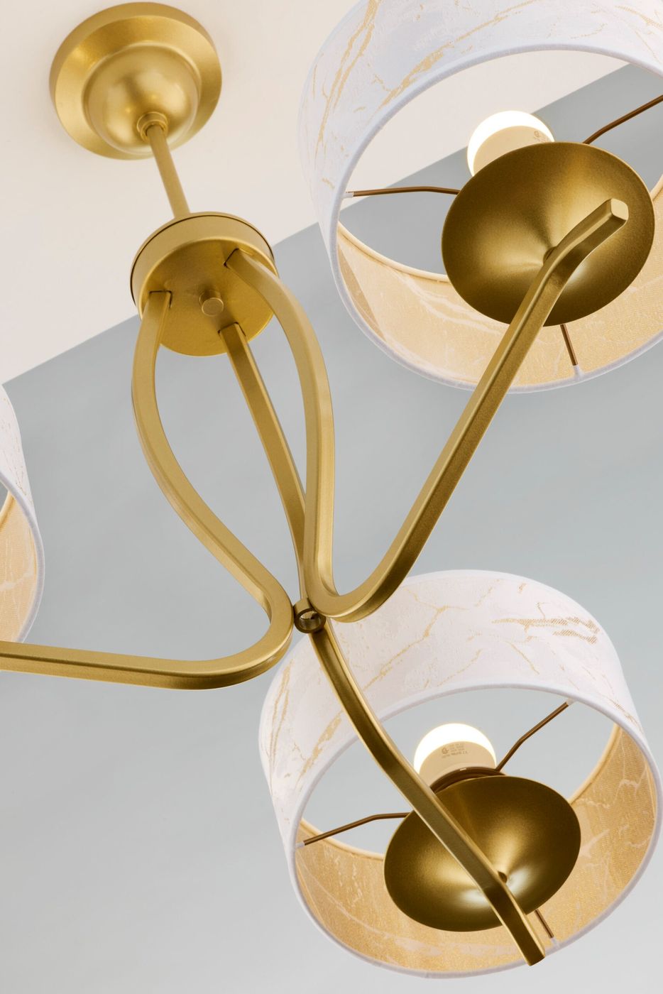 Żyrandol Kaszmir w kolorze złotym z trzema kloszami w białym odcieniu z delikatnym wzorem. Ujęcie z dołu, pokazujące złote ramiona i oprawki E27. Klosze są okrągłe, z widocznymi źródłami światła w środku. Idealny do nowoczesnych wnętrz.