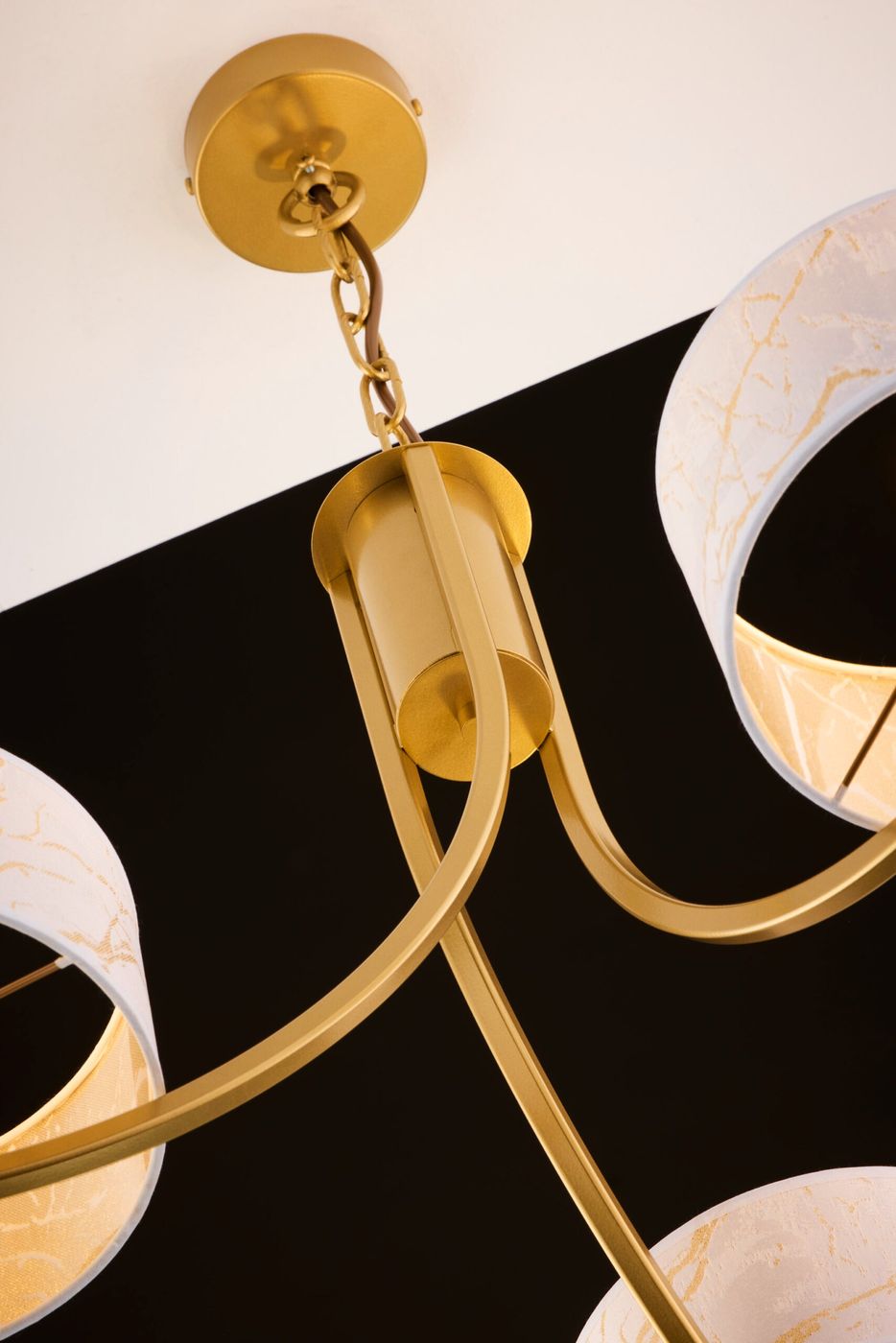Arthur żyrandol w kolorze złotym z białymi abażurami, z trzema miejscami na źródła światła E27. Ujęcie z góry, ukazujące metalowy stelaż i detale łączeń. Abażury z delikatnym wzorem, umieszczone na okrągłych mocowaniach.