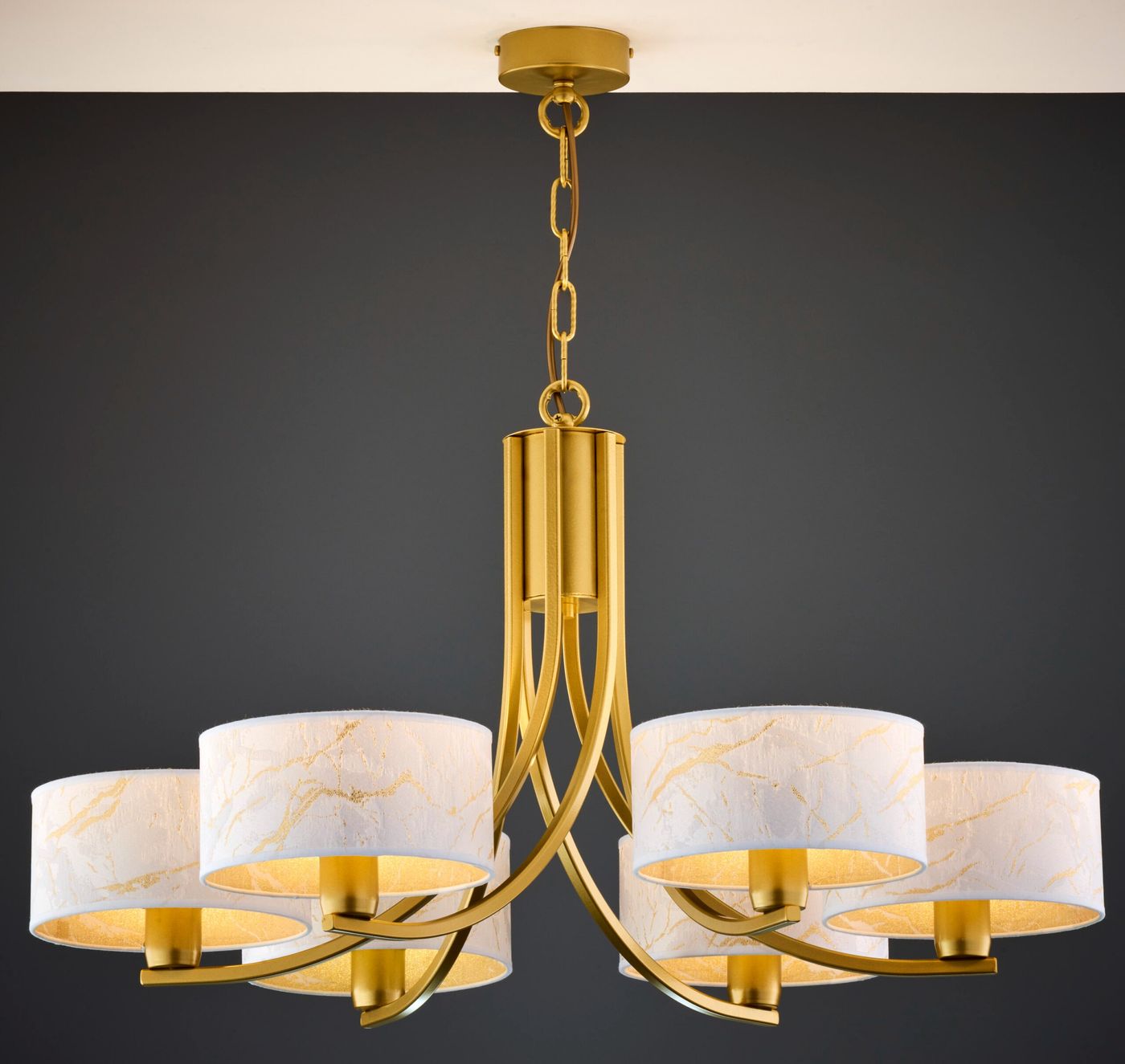 Żyrandol Arthur w kolorze złotym z białymi abażurami, przeznaczony dla sześciu żarówek E27. Posiada elegancką, zaokrągloną formę z metalowymi ramionami, zawieszony na łańcuchu. Tło ciemne, podkreślające jego styl.