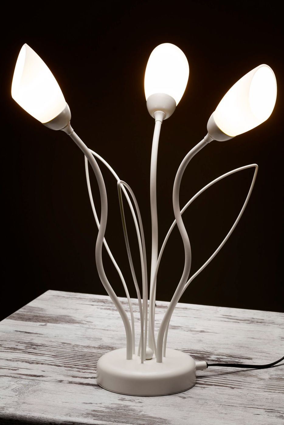 Lampa stołowa Gloriosa w kolorze piaskowo-szarym z trzema źródłami światła G9. Kształt przypominający kwiaty z białymi kloszami na cienkich, giętkich ramionach. Umieszczona na okrągłej podstawie z przewodem zasilającym.