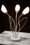 Lampa stołowa Gloriosa w kolorze piaskowo-szarym z trzema źródłami światła G9. Kształt przypominający kwiaty z białymi kloszami na cienkich, giętkich ramionach. Umieszczona na okrągłej podstawie z przewodem zasilającym.