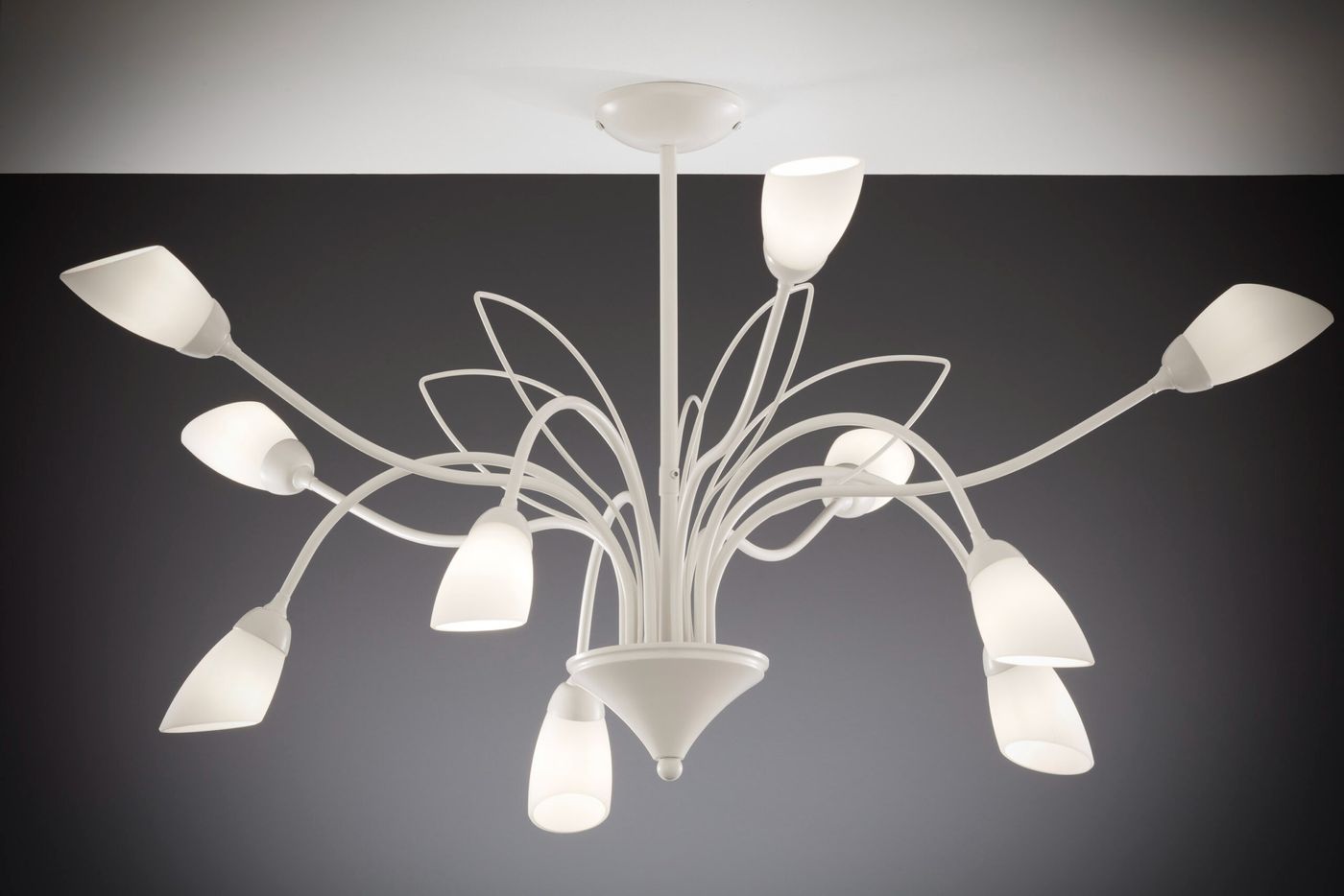 Żyrandol Gloriosa w kolorze piaskowo-szarym z 10 kloszami G9. Ma smukłą, białą konstrukcję z wygiętymi ramionami, prezentowany z góry, zawieszony na suficie. Klosze mają kształt delikatnych kielichów, co podkreśla elegancki design.