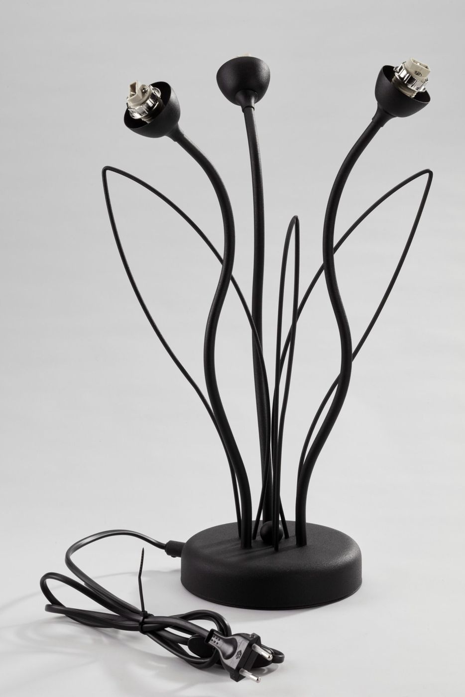 Gloriosa lampa stołowa z trzema gniazdami G9, czarna, z metalowymi, stylizowanymi ramionami przypominającymi kwiaty, z okrągłą podstawą i kablem z wtyczką widocznym z boku. Ujęcie z boku, na jasnym tle.