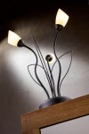 Lampa stołowa Gloriosa w kolorze czarnym z trzema kloszami w kształcie kwiatu, wykonana z metalu. Ujęcie z boku, pokazujące delikatne, wygięte ramiona oraz gładką podstawę. Klosze w kolorze białym, emitujące ciepłe światło.