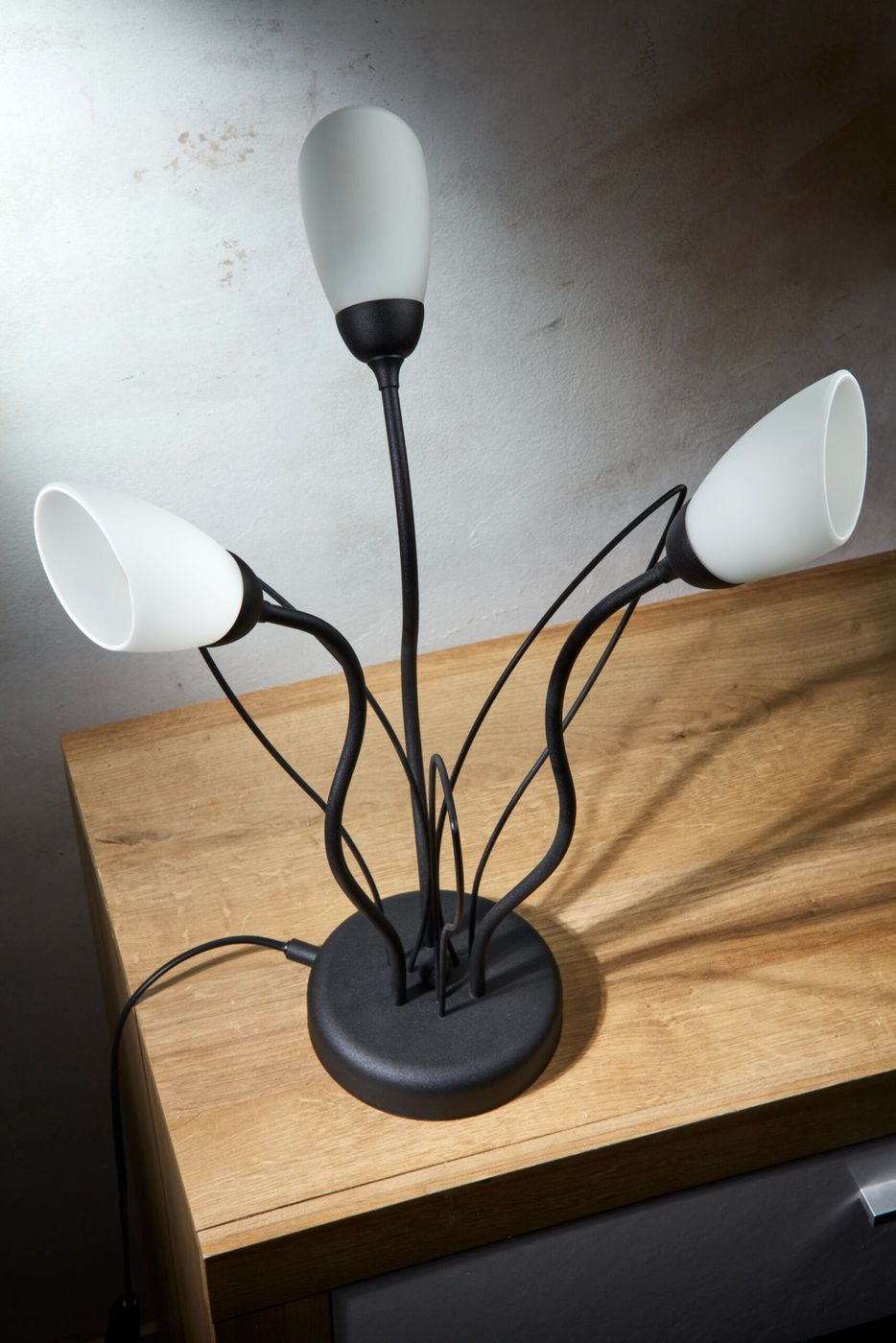 Lampa stołowa Gloriosa z trzema kloszami w kolorze białym, wykonana z czarnych metalowych elementów. Ułożona na drewnianym blacie, z widocznym kablem. Klosze mają elegancki kształt i są lekko zaokrąglone.