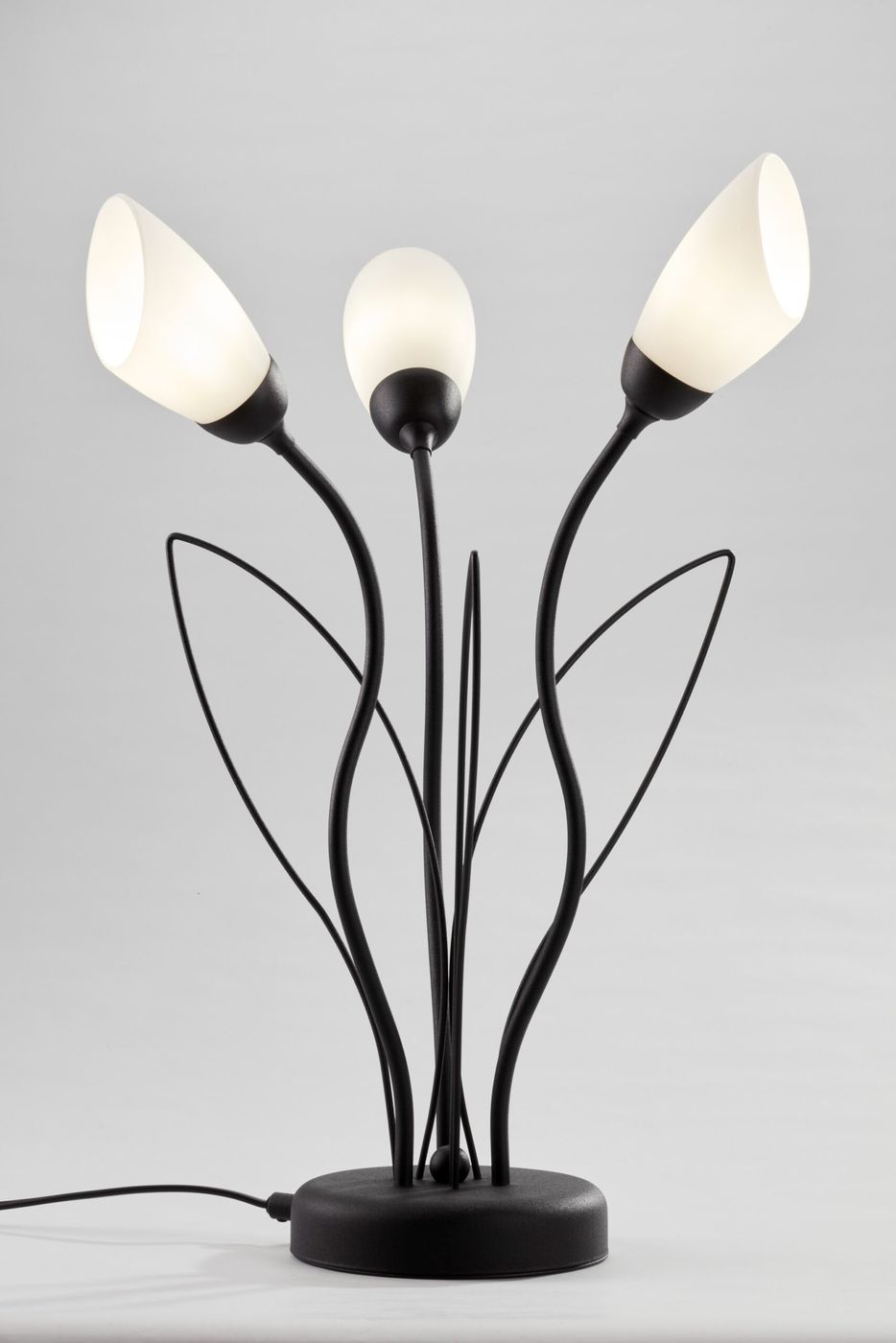 Lampa stołowa Gloriosa 3xG9 w czarnej kolorystyce, z trzema białymi kloszami w kształcie kwiatów. Metalowa podstawa z delikatnymi, zakrzywionymi ramionami. Produkt ustawiony na płaskiej powierzchni, z widocznym przewodem zasilającym.