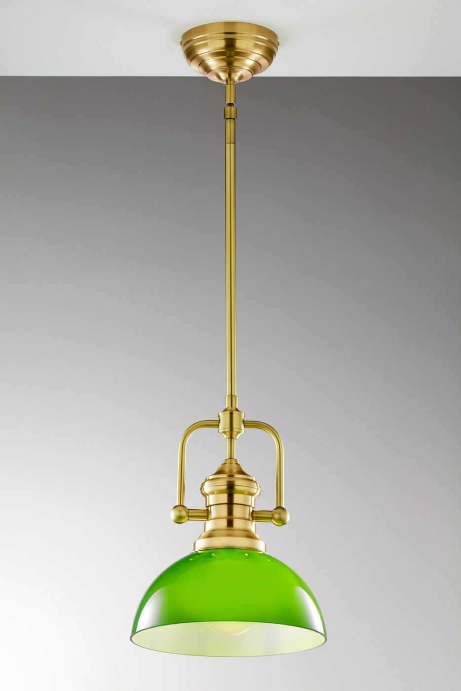 Lampa wisząca Platino z mosiądzu szczotkowanego, z zielonym kloszem. Ujęcie frontalne, zawieszona na cienkim drucie. Klosz ma okrągły kształt i jest lekko przezroczysty, eksponując źródło światła wewnątrz. Tło w odcieniach szarości.