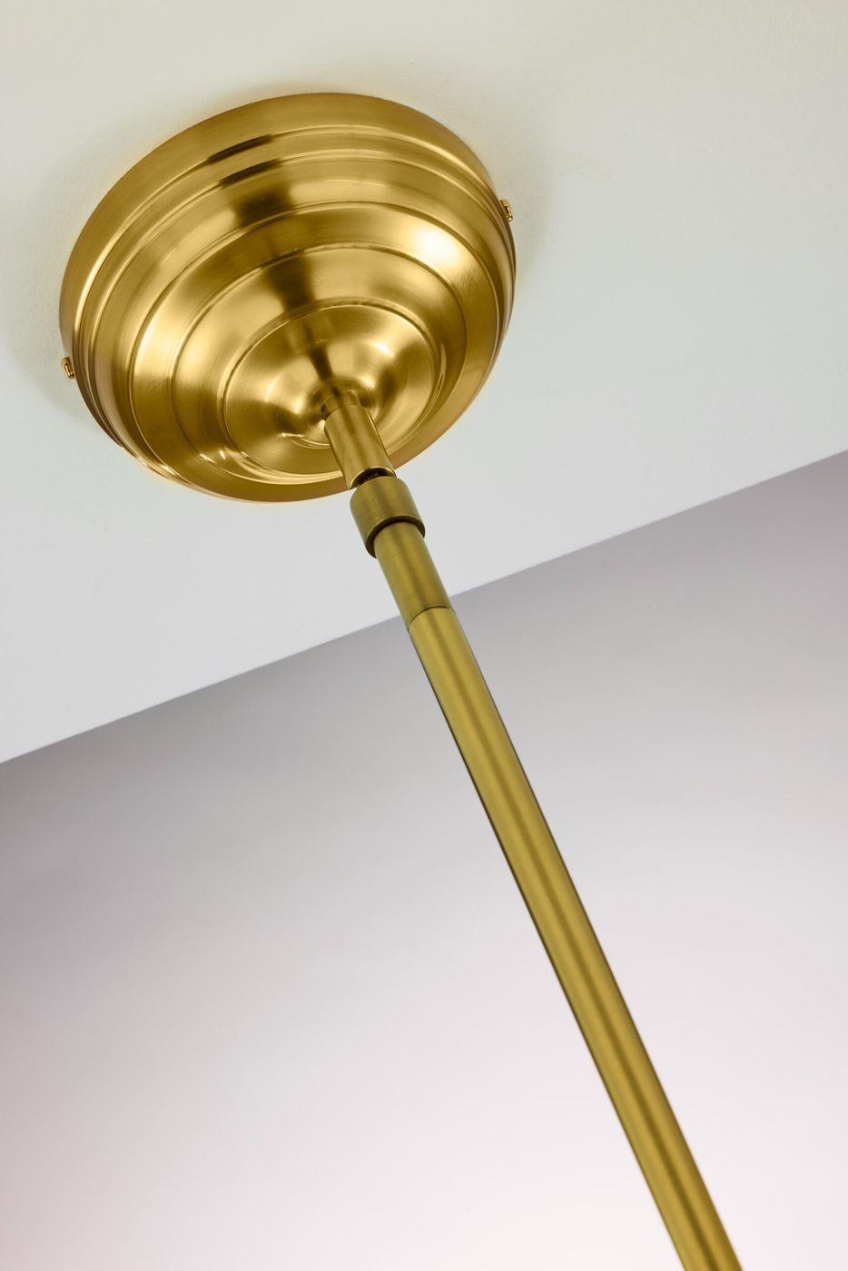 Lampa wisząca Platino w kolorze mosiądzu szczotkowanego, z pojedynczym gniazdem E27. Ujęcie z dołu pokazuje okrągły, dekoracyjny element montażowy oraz długi, prosty trzon. Tło w jasnym kolorze, co podkreśla elegancję lampy.