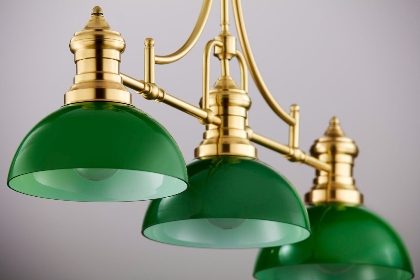 Lampa wisząca Platino z trzema kloszami w zielonym kolorze, wykonana z mosiądzu szczotkowanego. Klosze mają zaokrąglony kształt, a całość zawieszona jest na eleganckiej konstrukcji. Ujęcie z przodu, z bliska, ukazujące detale wykończenia.