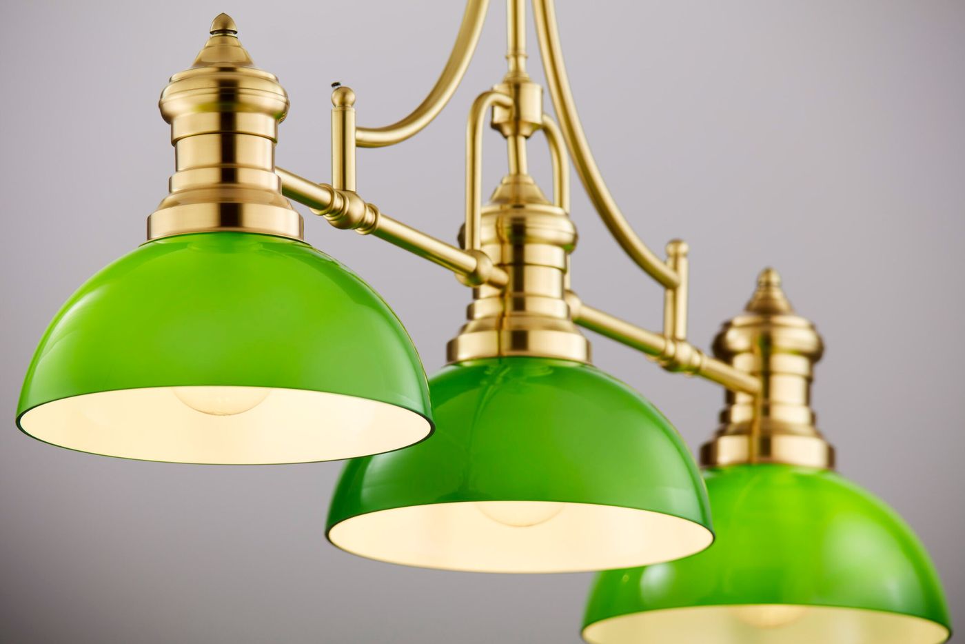 Lampa wisząca Platino z trzema kloszami w kolorze zielonym, wykonana z mosiądzu szczotkowanego. Ujęcie z przodu, klosze mają zaokrąglony kształt i białe wnętrze. Elementy metalowe mają złoty odcień, całość zawieszona na eleganckiej konstrukcji.