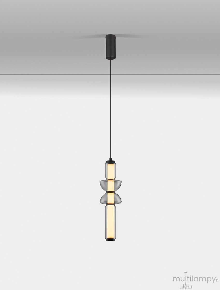 Lampa wisząca Glorica w kolorze czarnym, z jedną żarówką LED 15W. Wysoka, smukła konstrukcja z przezroczystymi elementami, w formie cylindrycznej z dekoracyjnymi detalami. Zawieszona na cienkim, czarnym przewodzie, na jasnym tle.