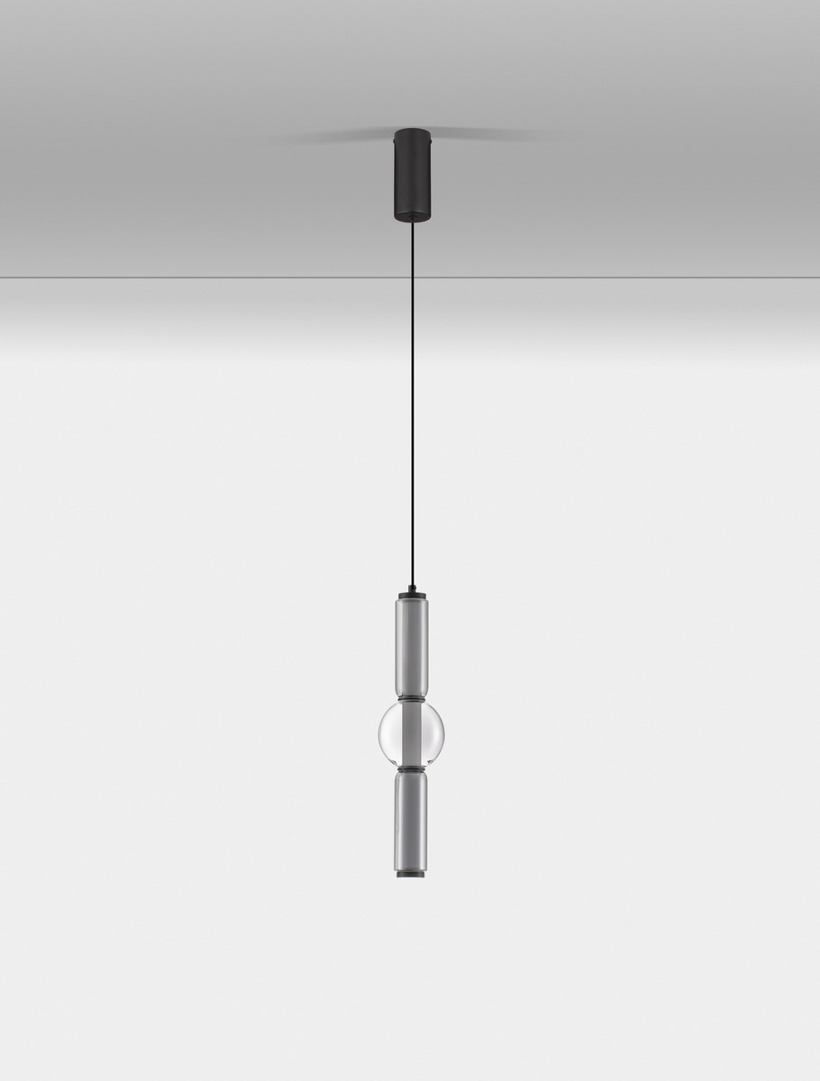 Lampa wisząca Glorica w kolorze czarnym, z jednym źródłem światła LED 15W. Posiada cylindryczny kształt z przezroczystą kulą w centralnej części. Zawieszona na czarnym przewodzie, montowana do sufitu. Tło białe, minimalistyczne.