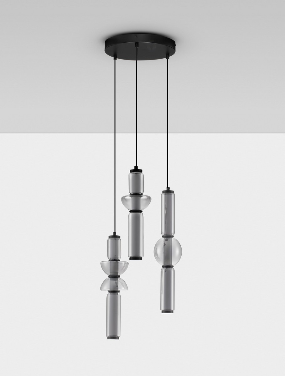 Lampa wisząca Glorica w kolorze czarnym, z trzema źródłami światła LED o mocy 37W. Posiada różne kształty kloszy: cylindryczny, okrągły i stożkowy, zawieszona na czarnych linkach, montowana do sufitu. Tło proste, jasne.