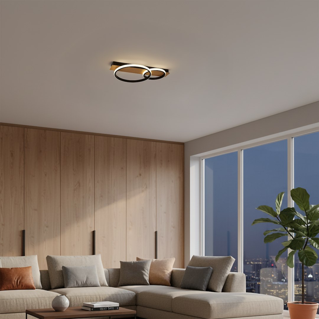 Czarna lampa sufitowa LED Rowena o mocy 14W i barwie światła 4000K, z dwoma okrągłymi pierścieniami. Zawieszona na gładkim suficie w nowoczesnym wnętrzu, w tle drewniana ściana i aranżacja z kanapą oraz dużymi oknami.